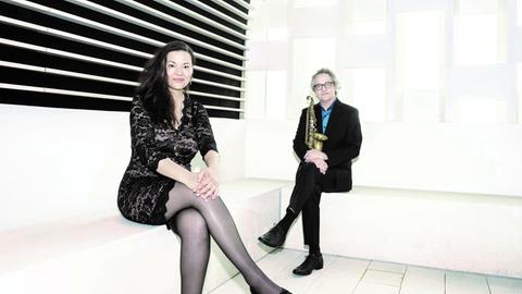 In der Ecke eines weißen Raumes sitzt im Vordergrund eine Frau im kurzen schwarzen Kleid, rechts im Hintergrund ein Mann im schwarzen Anzug und blauen Hemd. Beide lächeln.