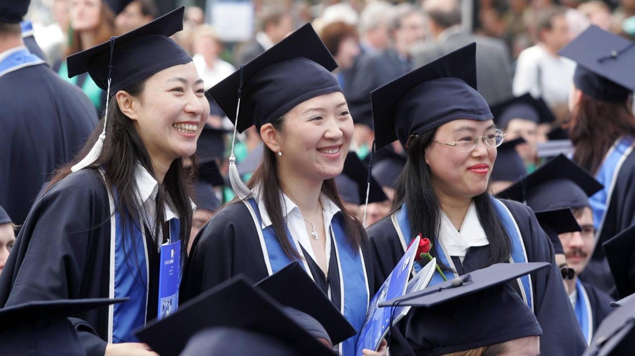 Drei asiatische Uni-Absolventinnen in Talar und Hut lächeln in eine Kamera.