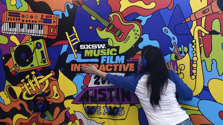 Eine Frau mit Kopfhörern auf den Ohren steht mit dem Rücken zum Betrachter vor einer bunt bemalten Wand auf der die Begriffe "SXSW", "Music", "Film", Interactive", "2018" und "Austin" zu lesen sind