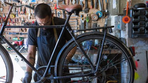 In einer Werkstatt mit einer Wand voller Werkzeuge wird ein altes Fahrrad von einem Mann mit Schürze repariert.
