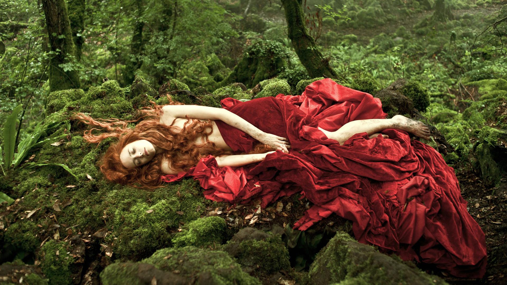 Stacey Martin liegt in roten Stoff eingehüllt auf einer Moosfläche in einem Wald.