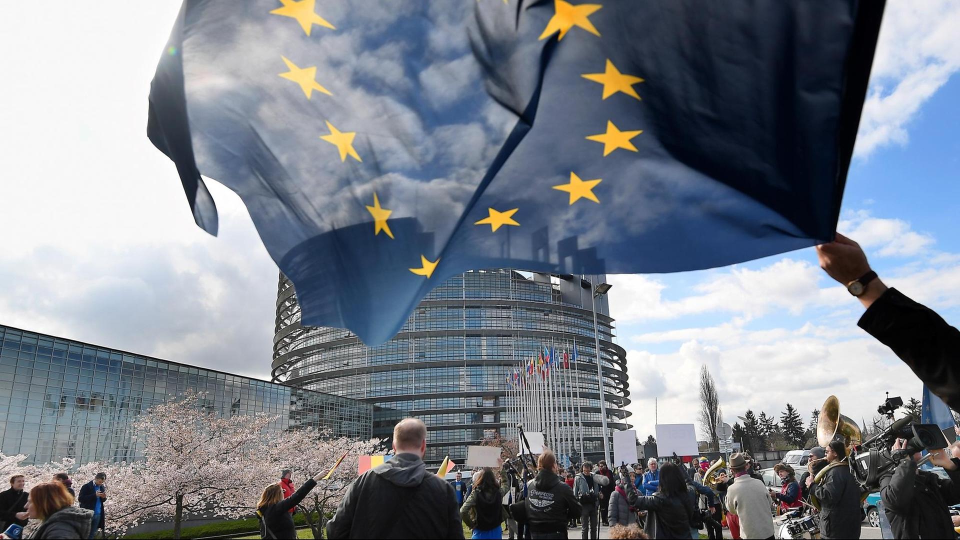Menschen stehen vor dem runden Parlamentsgebäude in Straßburg. Vorne im Bild schwenkt jemand eine EU-Flagge.