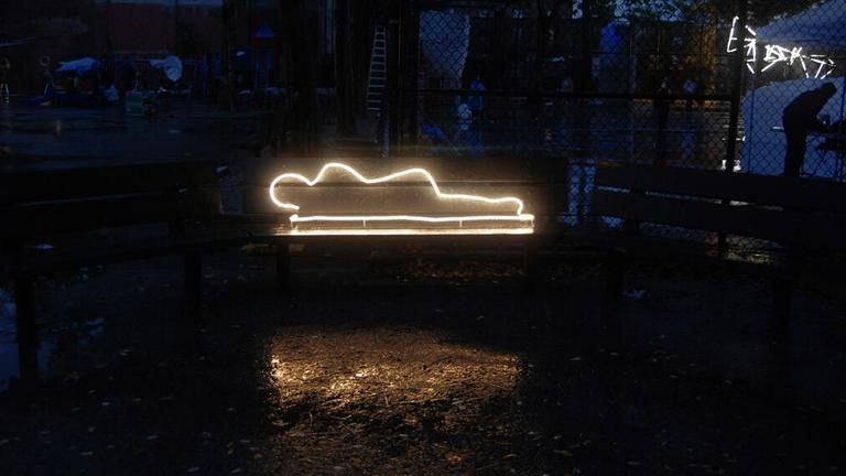Vor einem dunklen Hintergrund ist eine Neonskulptur zu sehen: die leuchtende Silhouette einer obdachlosen Person, die auf einer Matratze liegt
