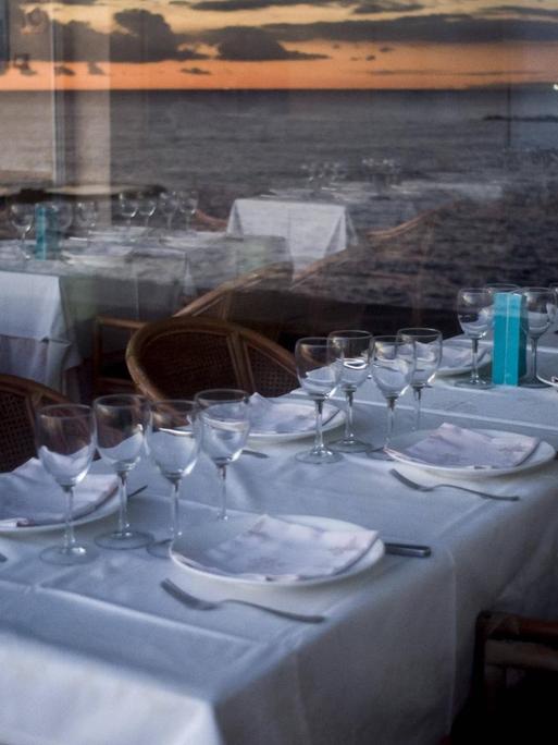 Leere Tische in einem Restaurant, in den Fensterscheiben spiegelt sich die Küste.