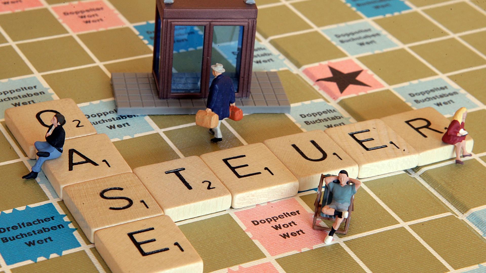 Eine Miniaturfigur trägt zwei Geldkoffer auf einem Scrabble-Spiel in eine Bank (Illustration), auf dem das Wort Steueroase liegt.