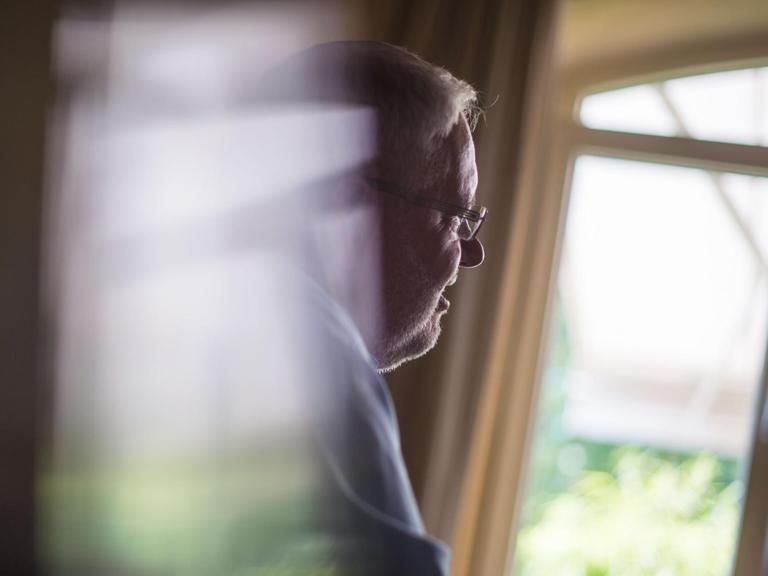 Auf dem Foto ist ein älterer Mann im Profil zu sehen, der aus einem Fenster schaut.