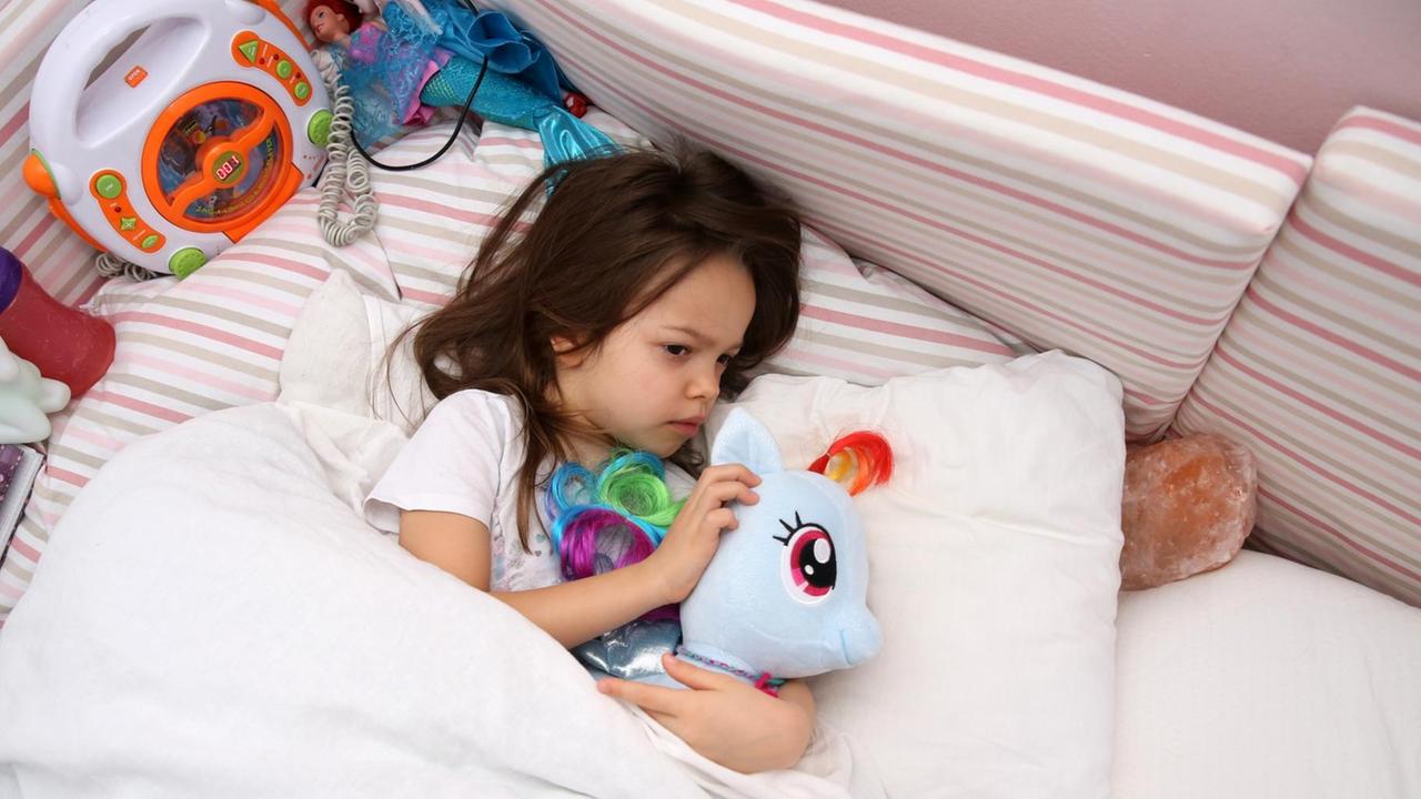 Ein kleines Maedchen liegt in einem Kinderzimmer und kuschelt mit ihrem Plüschtier (gestellte Szene).