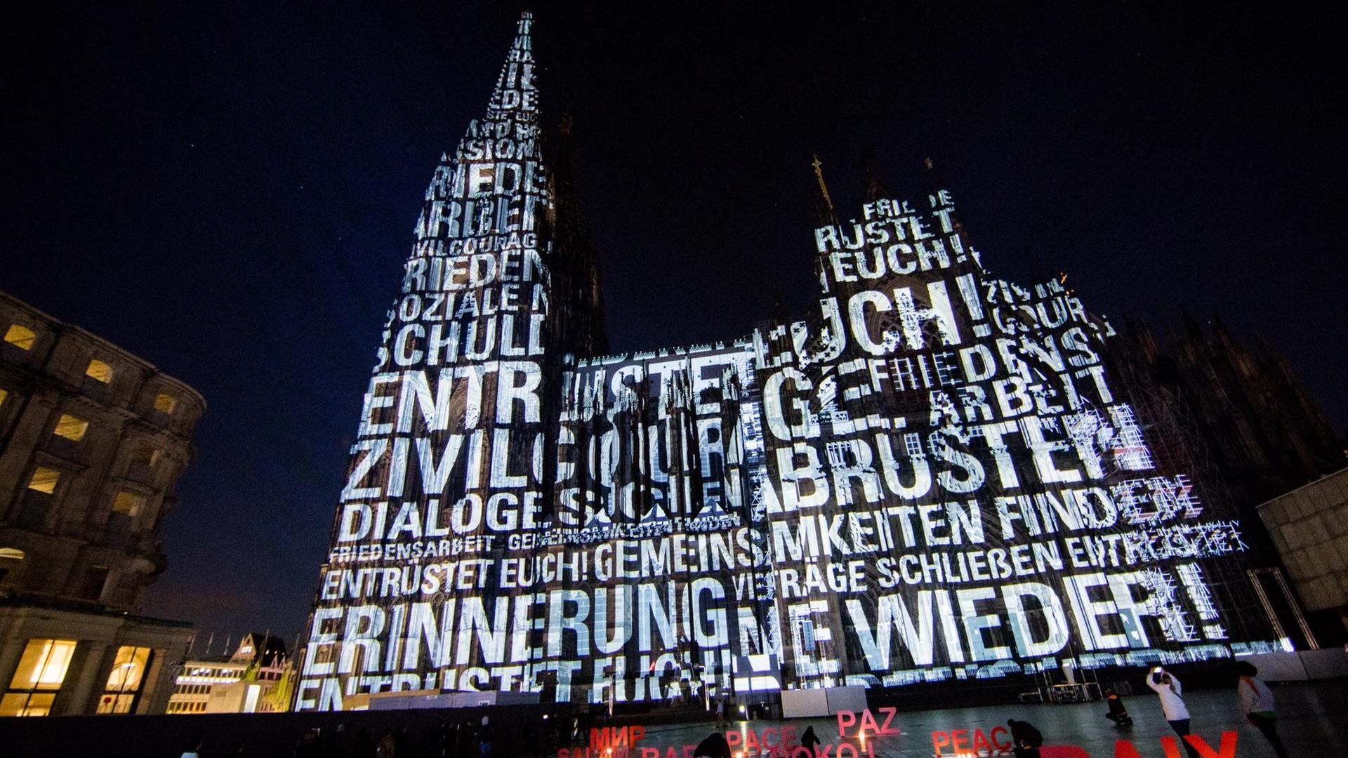 Das Foto zeigt den Kölner Dom. Auf der Wand von dem Dom sind große Wörter zu lesen.