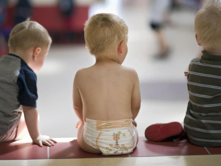 In einer Kindertageseinrichtung sitzen drei Kleinkinder auf einer Stufe
