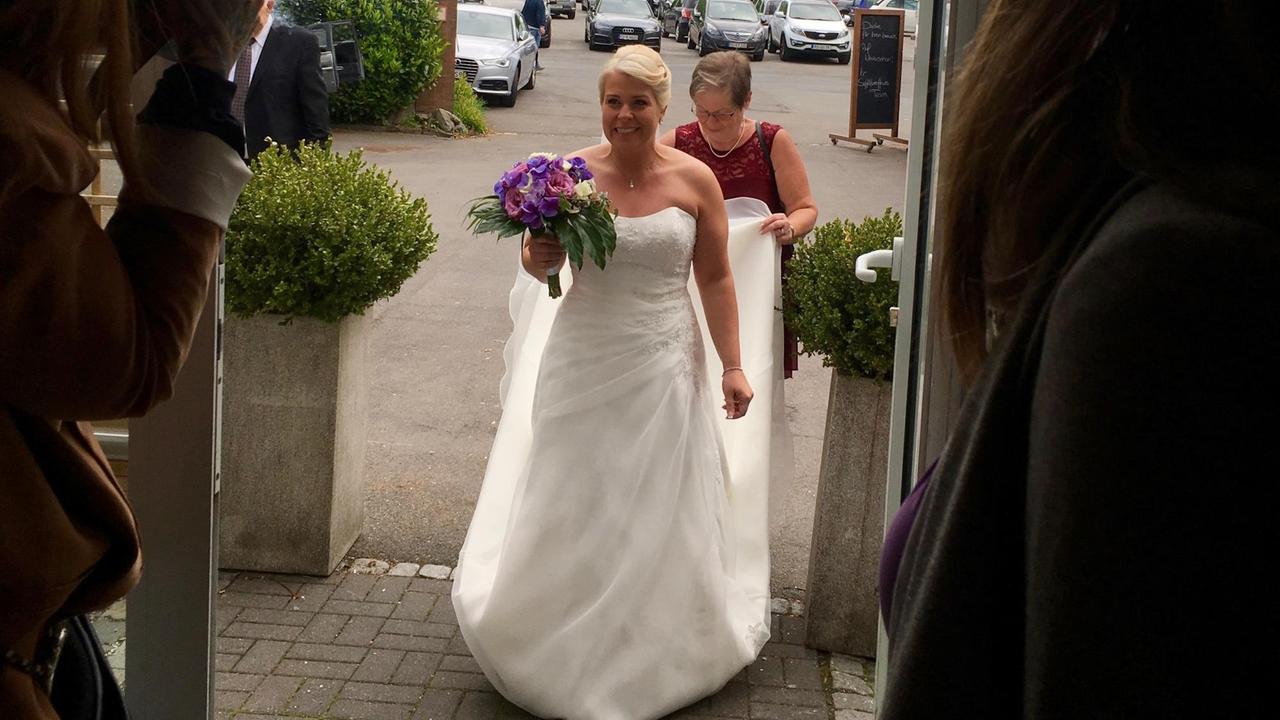 Eine Braut im weißen Kleid mit Schleppe tritt durch eine Tür 