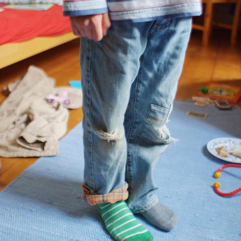 Ein kleiner Junge steht mit schmutziger Kleidung, zerrissenen Hosen und verschiedenen Strümpfen in einem unaufgeräumten Zimmer.