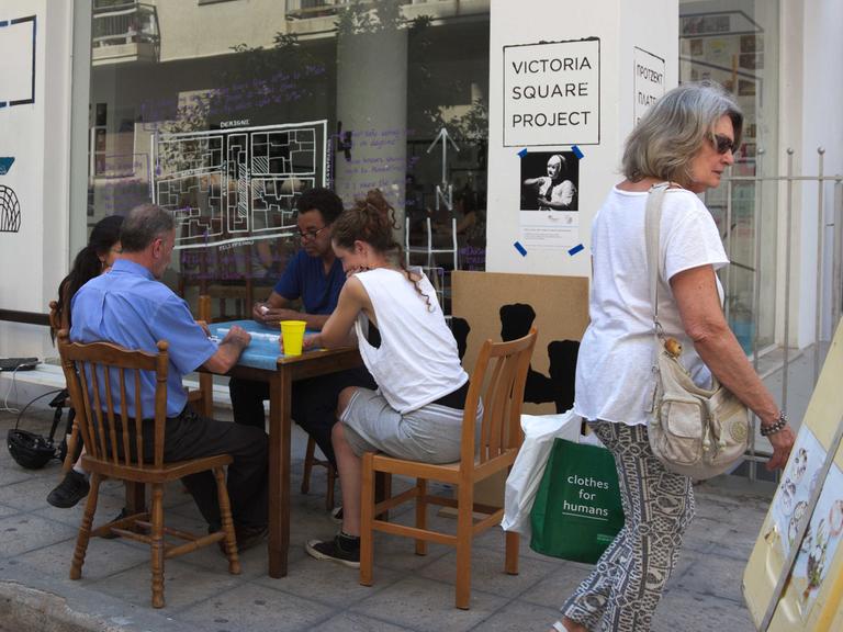 Riock Lowe sitzt mit drei Menschen um einen kleinen Holztisch am Rande einer Straße. Im Hintergrund ist ein verglastes Ladenblokal zu erkennen, das die Werkstatt für sein documenta 14-Projekt "Victoria Square Project" bildet.
