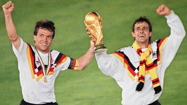 Lothar Matthäus und Pierre Littbarski halten den Pokal der Fußballweltmeisterschaft 1990 auf dem Rasen in die Höhe und winken.