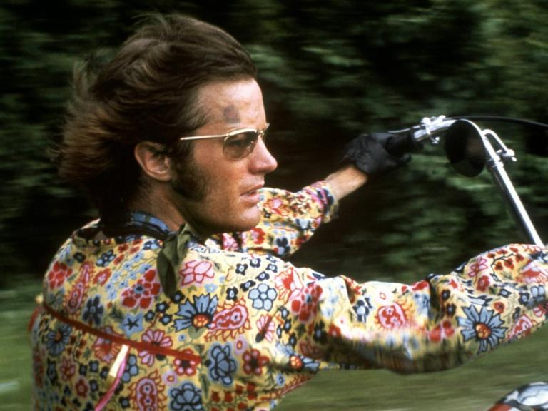 Peter Fonda mit buntem Blumenhemd und wehendem Haar in voller Fahrt auf seinem Motorrad im Film Easy Rider.