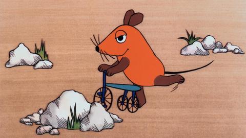 Ein Farbfoto zeigt die orangefarbene, offenbar erfreute Zeichentrick-Maus auf einem blauen Roller, umgeben von einigen weißen Felsen und kargen Grasbüscheln. Die erste Folge der "Sendung mit der Maus", ausgestrahlt am 7. März 1971 The Mouse Television: Die Sendung Mit Der Maus The Broadcast With The Mouse TV-Serie De 1971-, 07 March 1971 PUBLICATIONxINxGERxSUIxAUTxONLY Copyright: WdrxAFxArchivexMaryxEvans 12452915 editorial use only