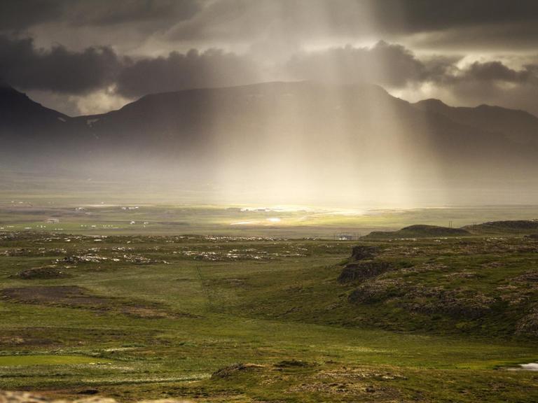 Sonnenlicht fällt durch die dichte, graue Wolkendecke auf die Landschaft Islands.