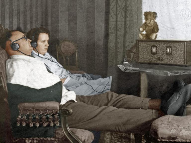 Ein Mann und eine Frau sitzen nebeneinander auf dem Sofa und hören mit Kopfhörern Radio, digital kolorierte Aufnahme aus dem Jahr 1928