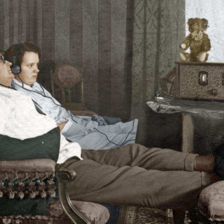 Ein Mann und eine Frau sitzen nebeneinander auf dem Sofa und hören mit Kopfhörern Radio, digital kolorierte Aufnahme aus dem Jahr 1928
