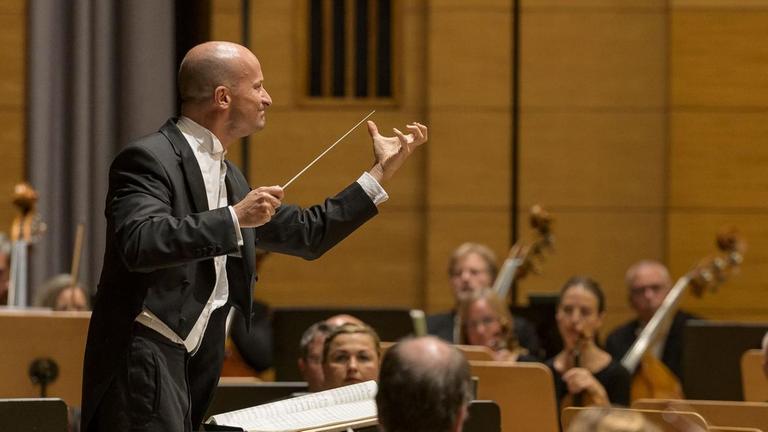 Ein Dirigent steht erhöht vor einem Orchester und feuert seine Musiker an, indem er seine Finger beinahe zu einer Faust gekrümmt hat.