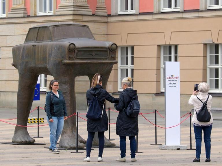 Passanten stehen vor der Skulptur eines Trabant auf Beinen.