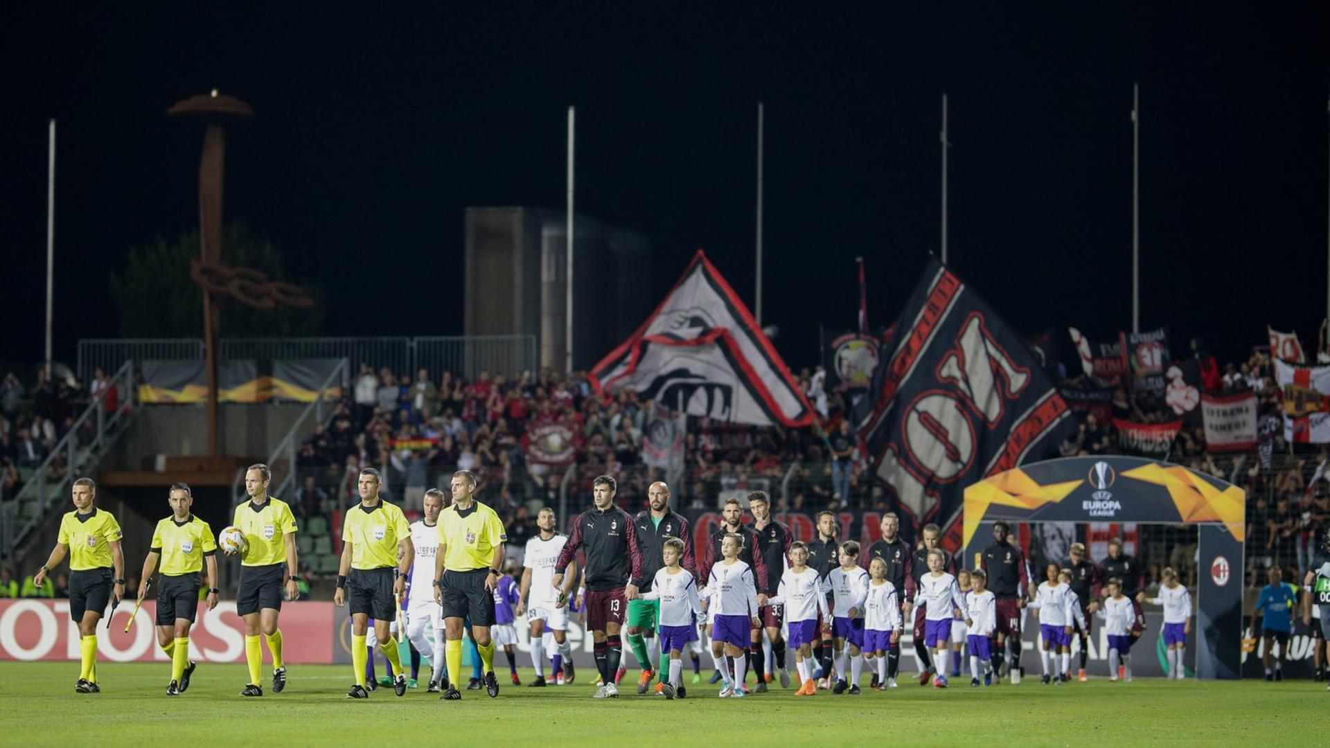 Fünf Schiedsrichter laufen vor den Mannschaften des F91 Düdelingen und des AC Mailand auf den Rasen eines Fußballstadions
