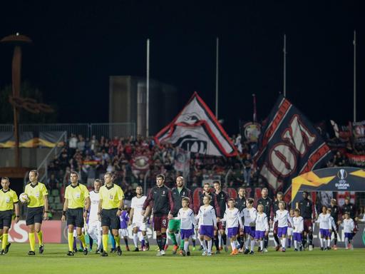 Fünf Schiedsrichter laufen vor den Mannschaften des F91 Düdelingen und des AC Mailand auf den Rasen eines Fußballstadions