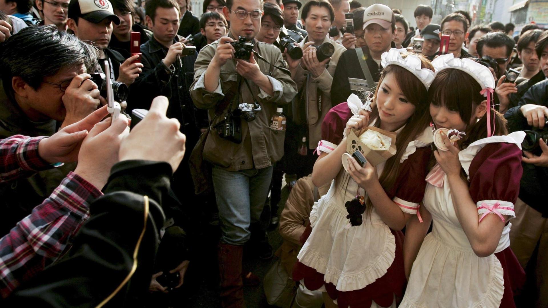 Zwei junge Japanerinnen, verkleidet als Anime-Charaktere, werden von Fans und Fotografen umringt.