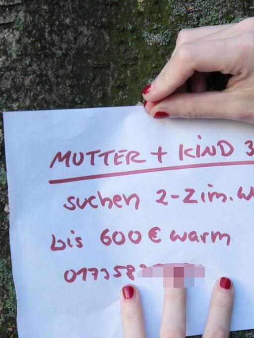 Die Hände einer jungen Frau, die auf Wohnungssuche ist, pint einen Zettel an einen Baum, auf dem steht: "Mutter und Kind suchen 2-3 Zimmer Wohnung".