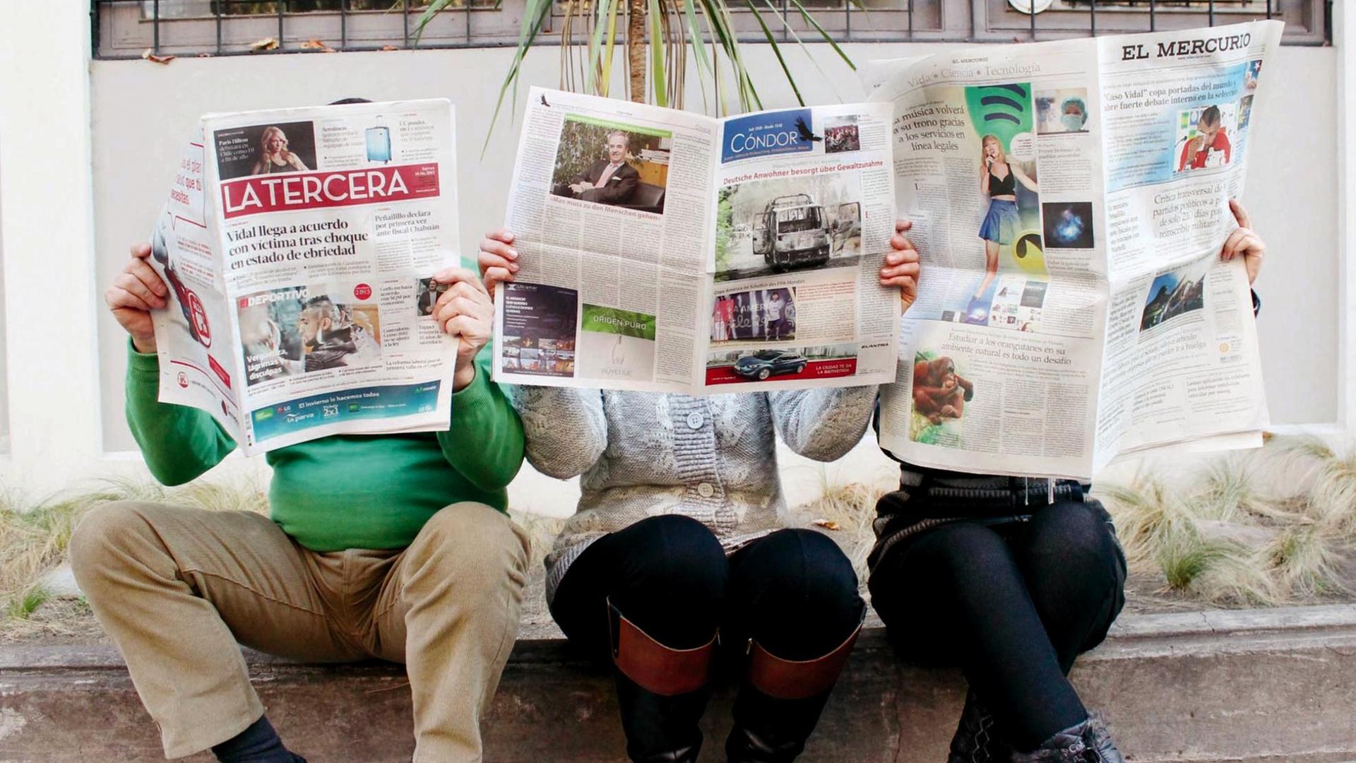 Auf einem Bürgersteig sitzen drei Personen, die Zeitungen lesen und deren Köpfe dahinter versteckt sind. Auf den Zeitungen sind die Titel zu lesen: "Latercera", "Condor" und "El Mercurio".