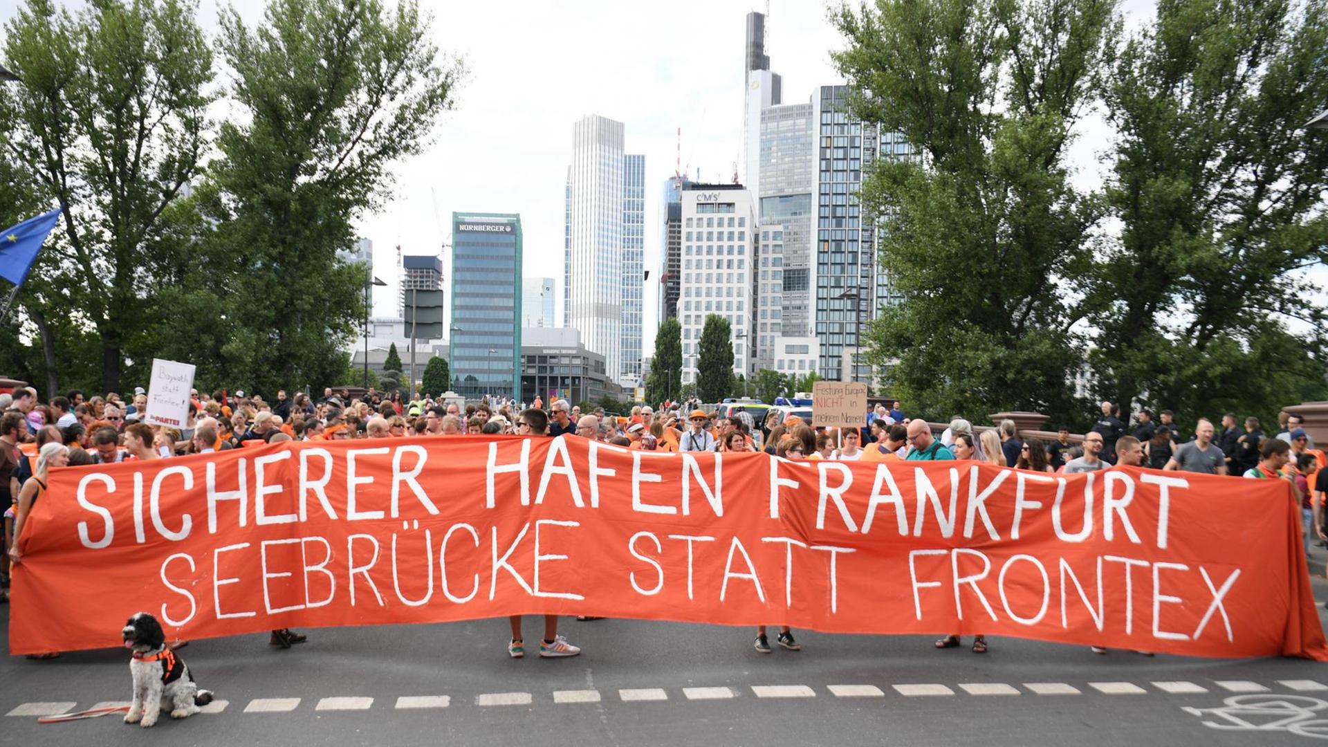 Demonstranten halten vor der Banken-Kulisse der Stadt ein Transparent mit der Aufschrift "Sicherer Hafen Frankfurt - Seebrücke statt Frontex" hoch.