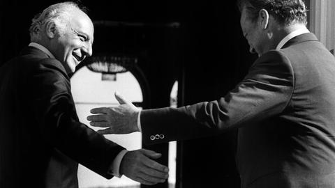 Schwarz-Weiß Foto von zwei Männern mittleren Alters, die sich freundlich zunicken, mit den Armen die Geste "Bitte, nach Ihnen" machen.