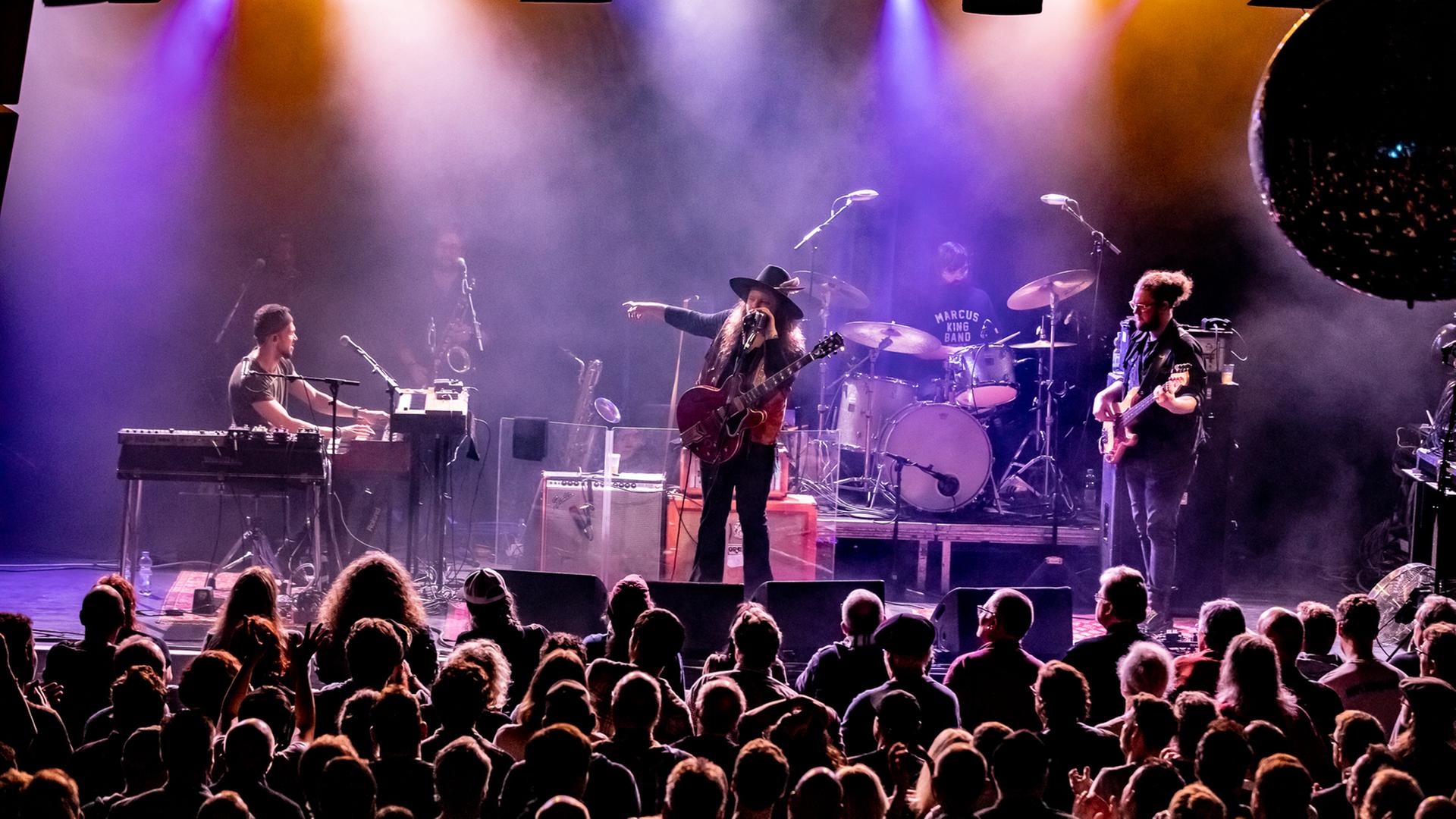 Sechs Männer stehen auf einer mit Nebel und Scheinwerferlicht gefluteten Bühne: der Sänger und Gitarrist in der Mitte zeigt auf jemanden rechts neben sich. Vor der Bühne steht Publikum.