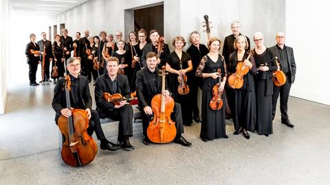 Viele Menschen mit Orchesterinstrumenten in der Hand stehen in einem weißen Raum.