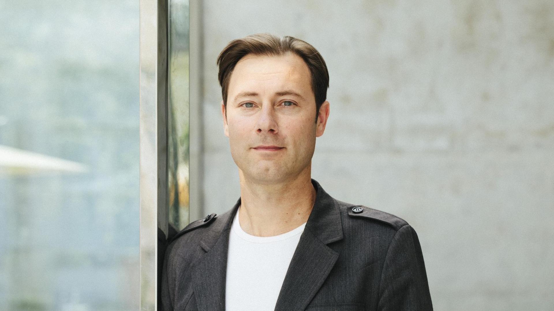 Thomas Biebricher, in weißem T-Shirt und grauem Sakko, blickt freundlich in die Kamera.