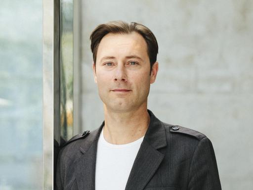 Thomas Biebricher, in weißem T-Shirt und grauem Sakko, blickt freundlich in die Kamera.