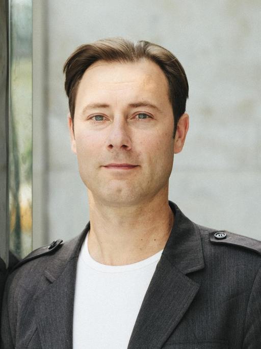 Thomas Biebricher, in weißem T-Shirt und grauem Sakko, blickt freundlich in die Kamera.