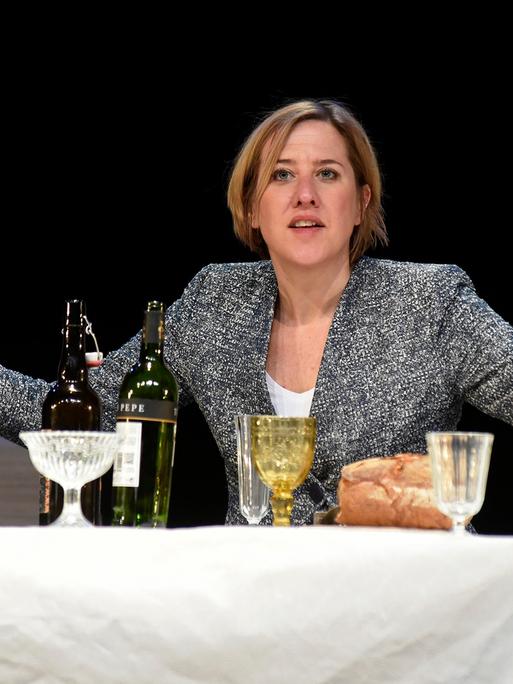Schauspielerin Silke Buchholz als Angela Merkel in einer Szene des Theaterstücks "Angela I" der Bremer Shakespeare Company. Sie sitzt an einer Tafel vor Weinflaschen und Gläsern.