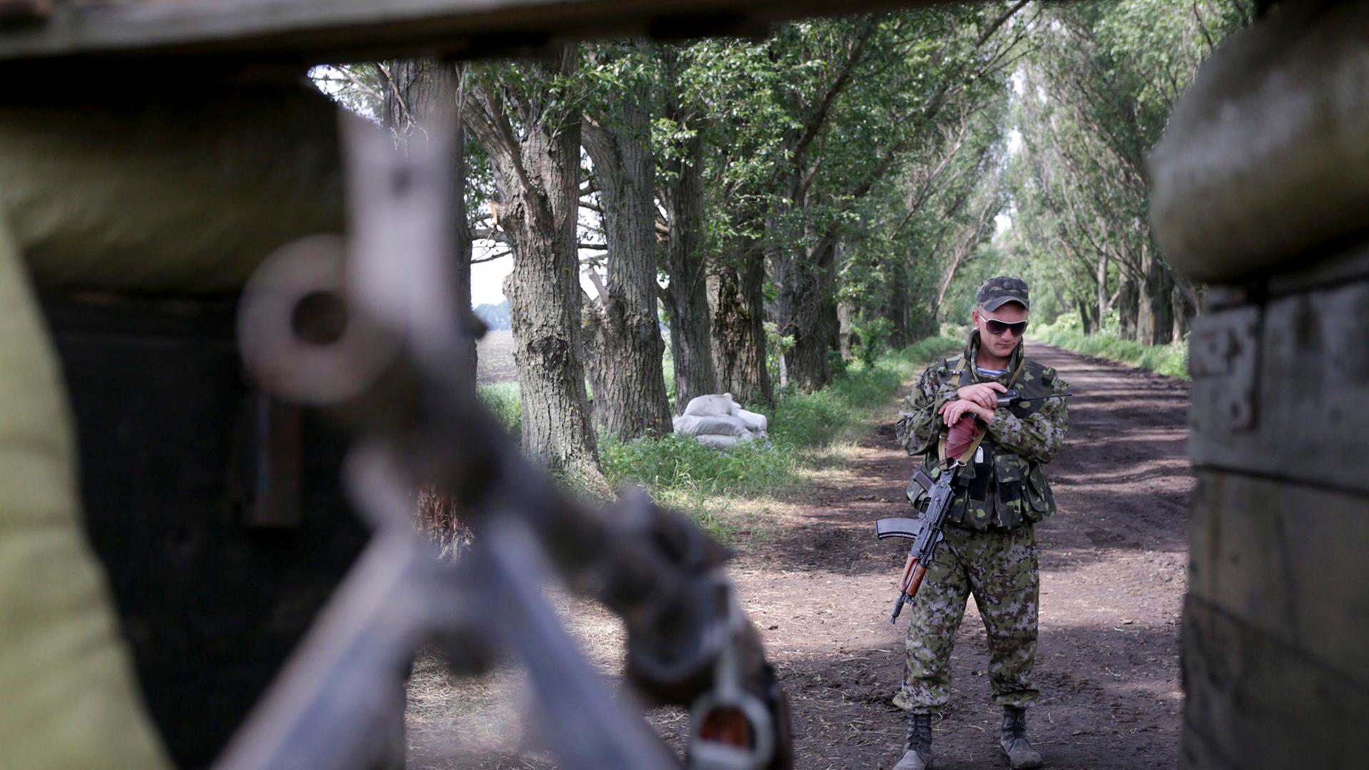 Ukrainischen Soldaten zielt mit einem Gewehr auf eine im Vordergrund ebenfalls mit einem Gewehr bewaffneten Person. Im Hintergrund ist eine Allee zu sehen.