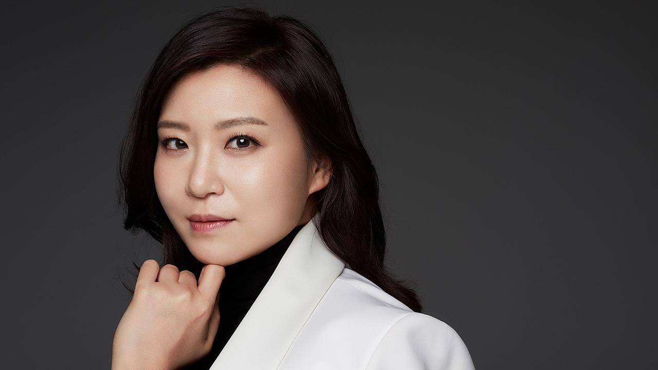 Die südkoreanische Dirigentin Shiyeon Sung trägt schwarzen Rollkragenpullover sowie ein weißes Jackett und blickt seitlich in die Kamera