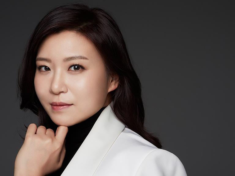 Die südkoreanische Dirigentin Shiyeon Sung trägt schwarzen Rollkragenpullover sowie ein weißes Jackett und blickt seitlich in die Kamera