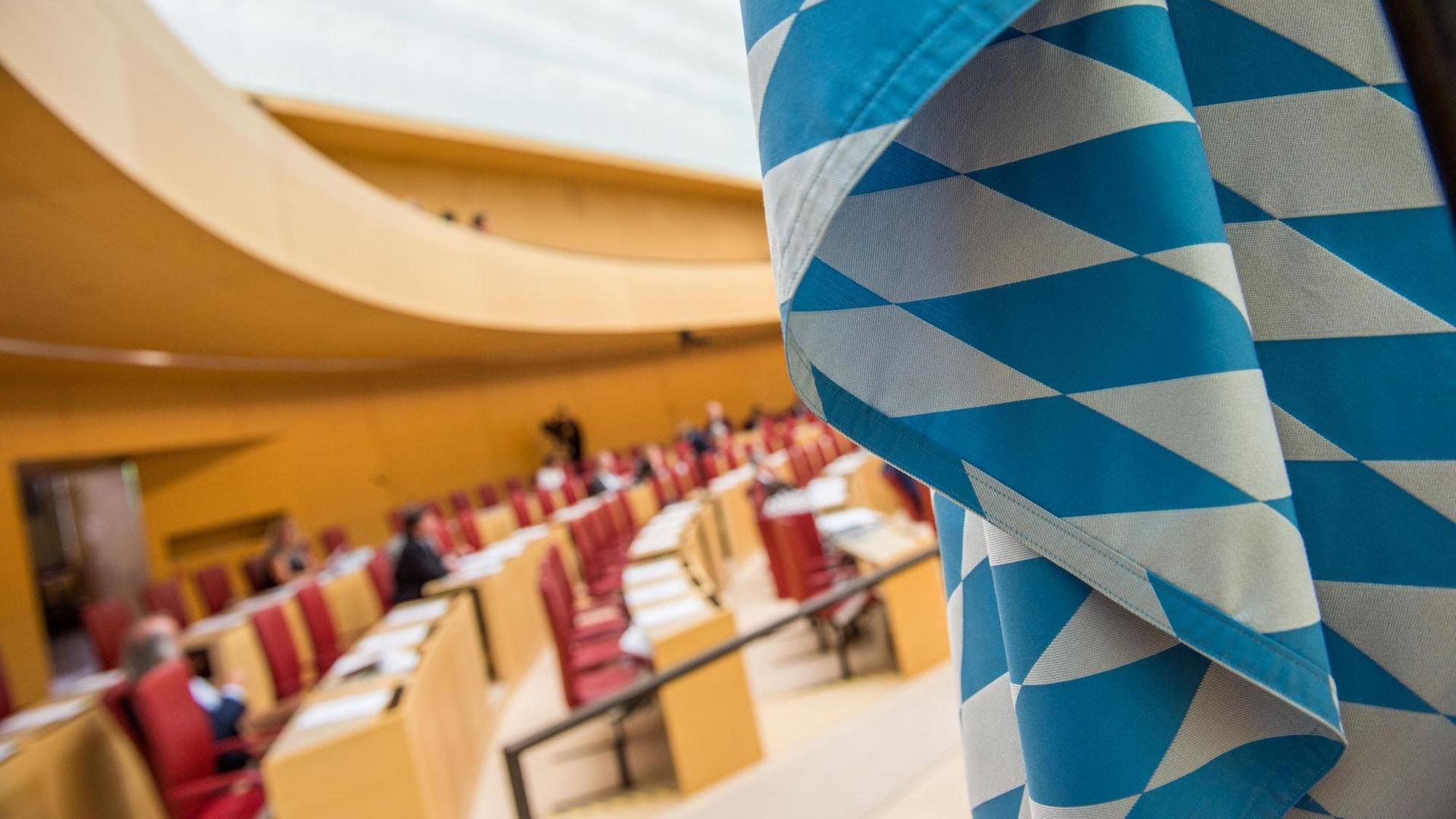 Man sieht einen Teil der blau-weißen Flagge scharf im Vordergrund und die teils besetzten Abgeordnetenbänke unscharf im Hintergrund.