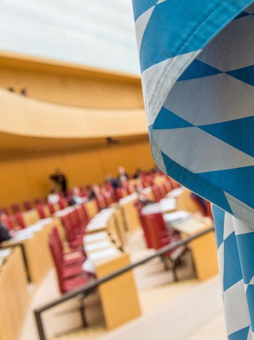 Man sieht einen Teil der blau-weißen Flagge scharf im Vordergrund und die teils besetzten Abgeordnetenbänke unscharf im Hintergrund.