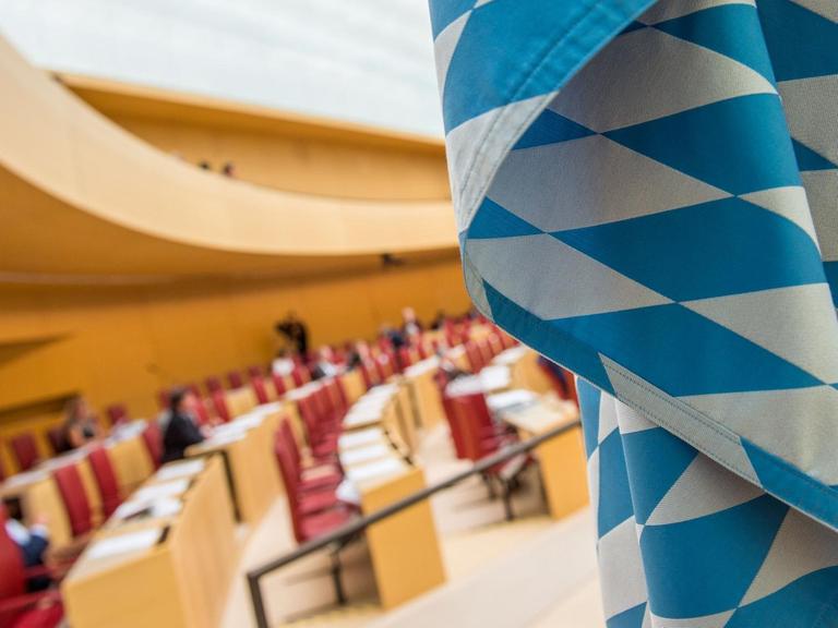 Man sieht einen Teil der blau-weißen Flagge scharf im Vordergrund und die teils besetzten Abgeordnetenbänke unscharf im Hintergrund.