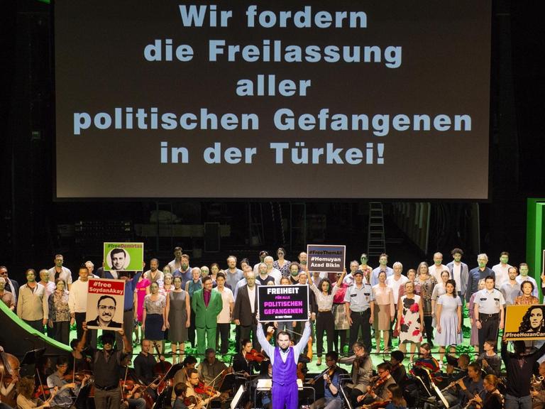 Auf einer riesigen Leinwand steht geschrieben: Wir fordern die Freilassung aller politischen Gefangenen in der Türkei! Davor stehen sehr viele Menschen mit grün bemalten Gesichtern und zum Teil mit Plakaten.