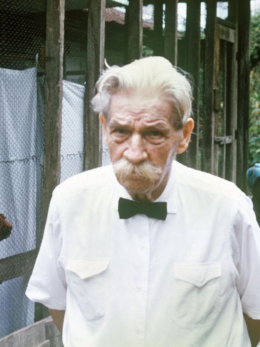 Älterer Mann mit Schnauzbart und einem weißen Hemd in einem afrikanischen Krankenhaus, im Hintergrund sitzen afrikanische Menschen 