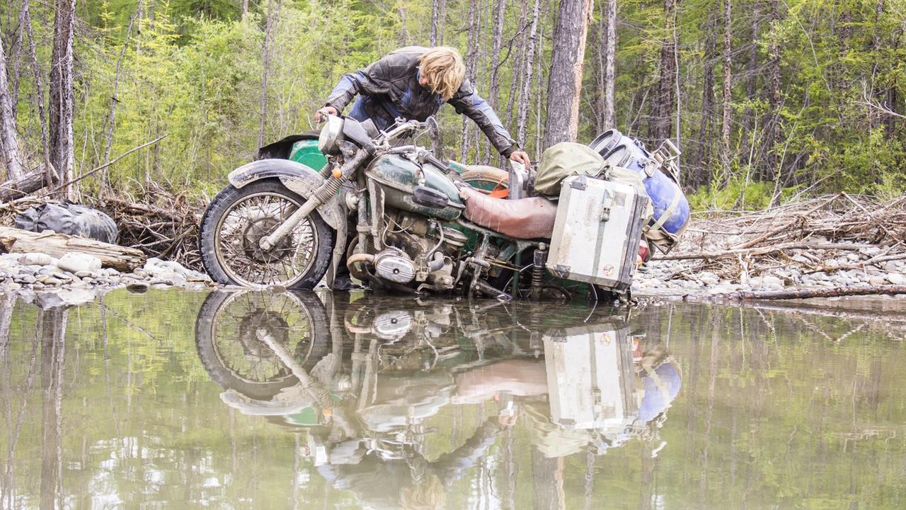 Ein Motorradfahrer kämpft sich mit seinem Motorrad durch ein großes, mit Wasser gefülltes Schlammloch. Das Motorrad ist schwer beladen.