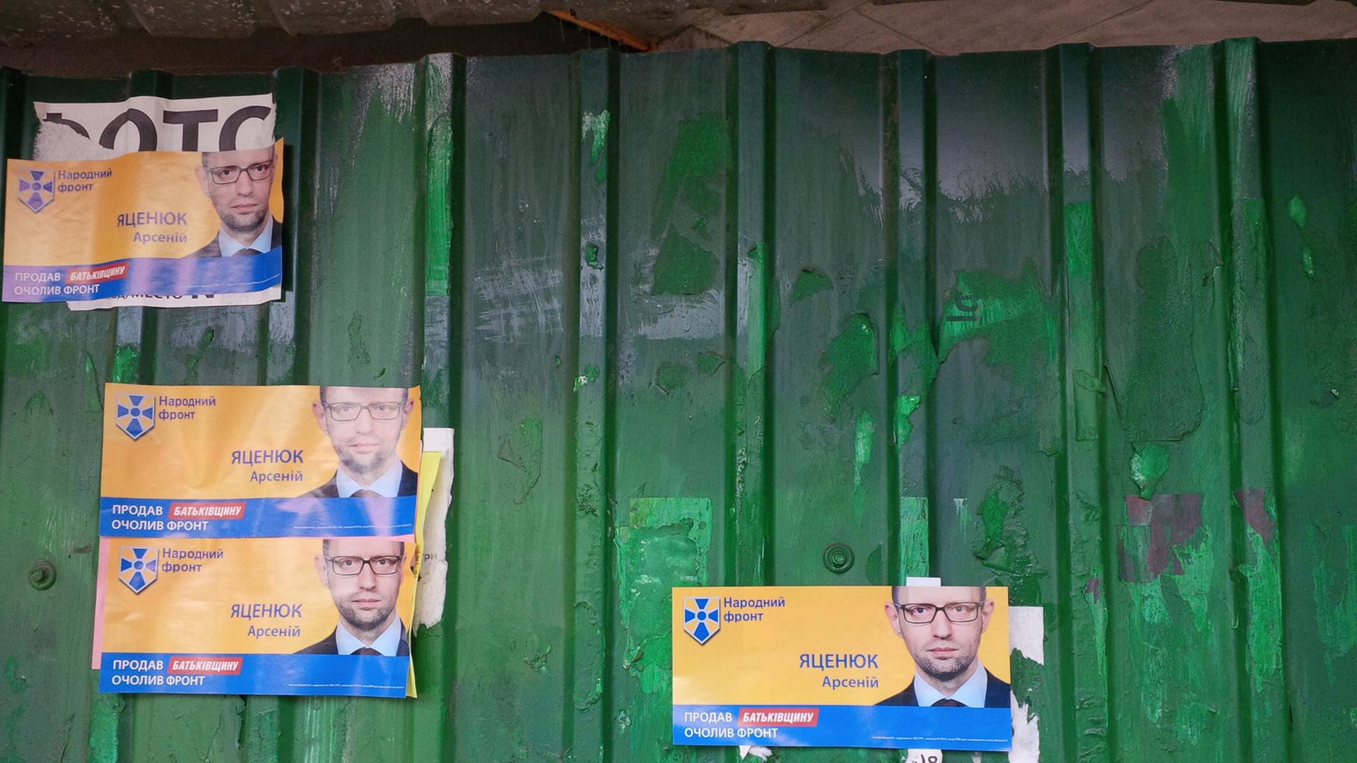 Mehrere kleine Plakate, auf denen der ukrainische Ministerpräsident Arsenij Jazenjuk zu sehen ist, kleben an einem grünen Zaun.