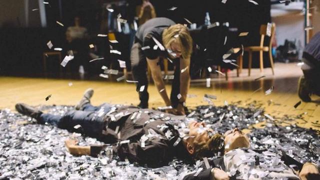 Szene aus dem Theaterstück "Cum-Ex Papers" im Lichthof Hamburg, Regie Hege Schmidt: Zwei Männer liegen auf dem Boden, in mitten von Konfetti-Streifen, ein dritter tritt dazu.