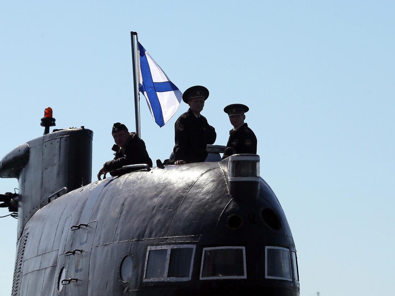 Auf einem U-Boot stehen drei Männer und es ist eine Flagge mit blauem Andreaskreuz auf weißem Grund zu sehen.
