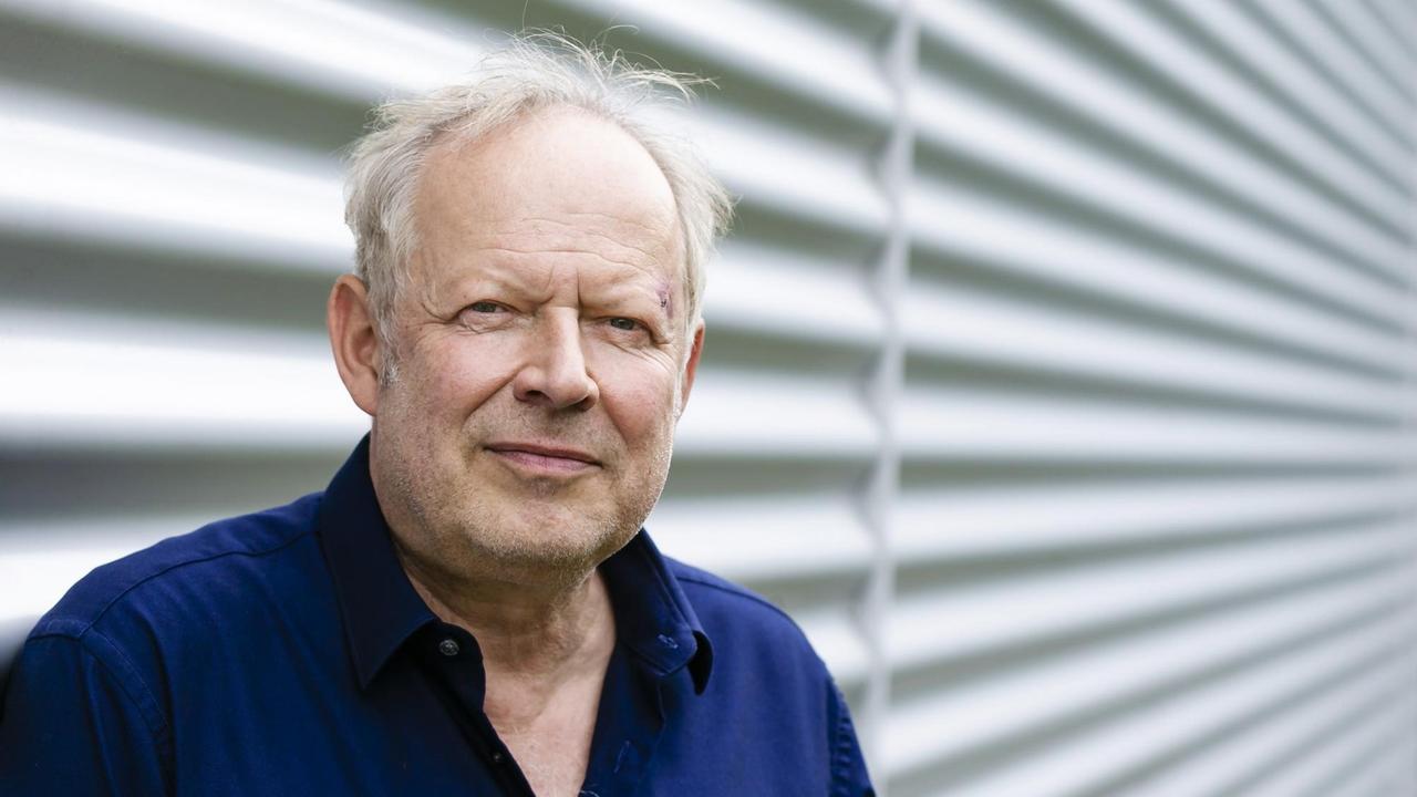 Porträt von Axel Milberg im blauen Hemd: Er steht lächelnd vor einer Wand aus Wellblech.